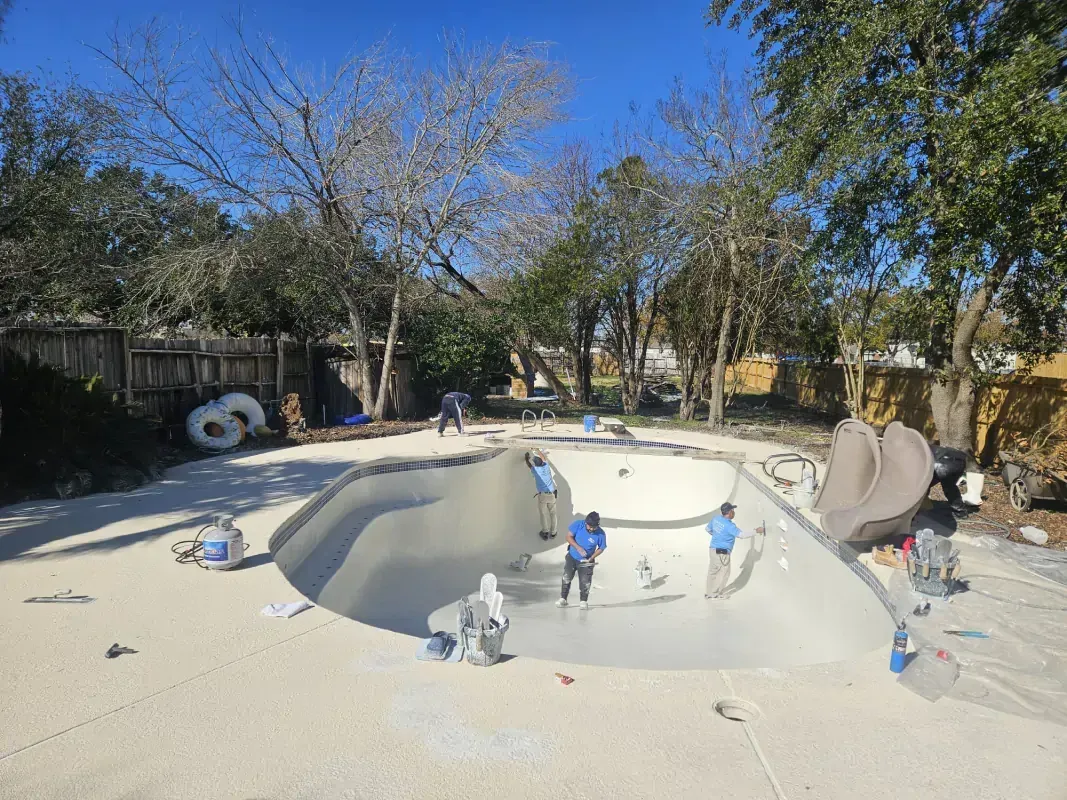 Two people working inside a partially constructed, light-colored swimming pool in a backyard.