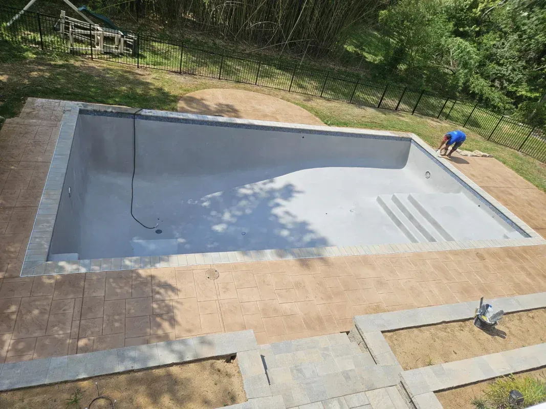 An empty, gray-lined rectangular swimming pool with stone coping, surrounded by a patio, with a person working nearby.