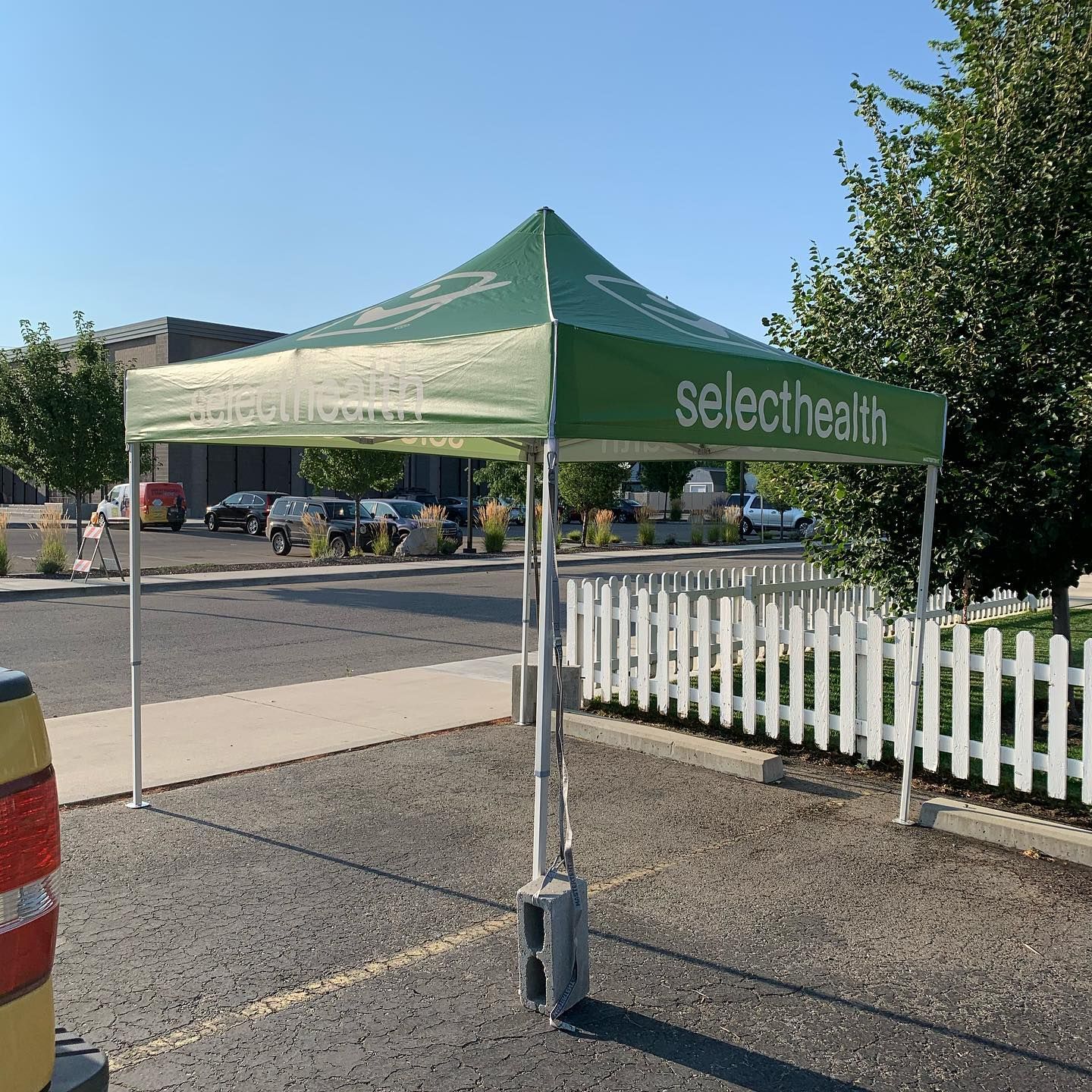 A green SelectHealth pop-up tent stands on an asphalt lot next to a white picket fence.