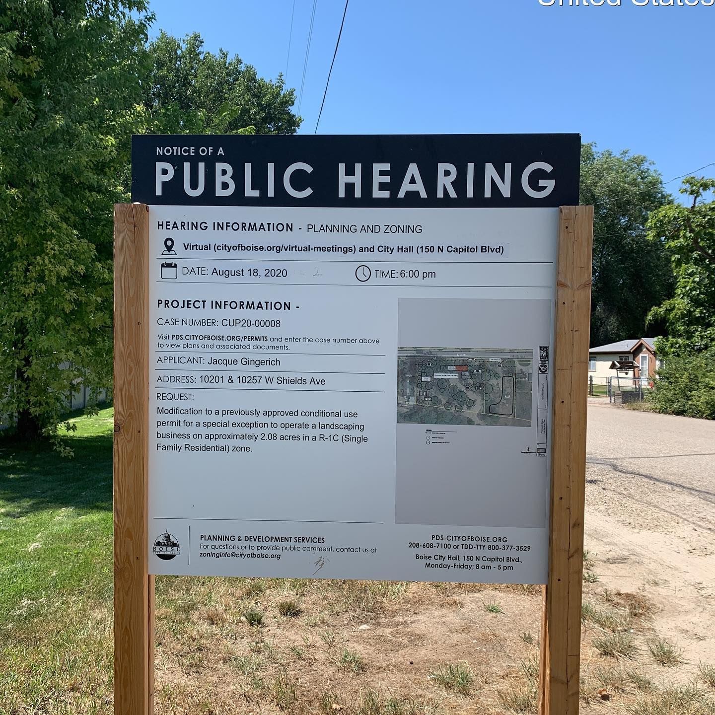 A Public Hearing notice sign on wooden posts, displaying project information and a site map against a background of trees.