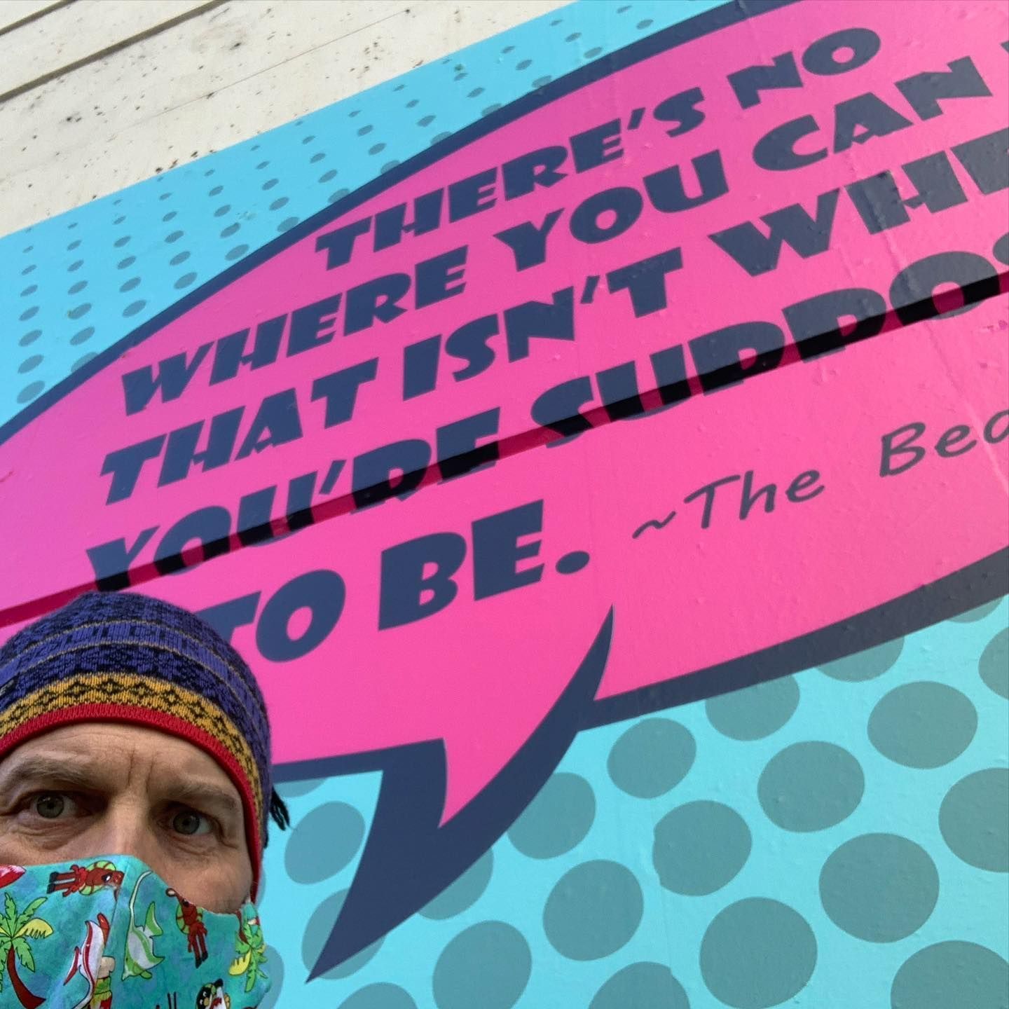 A person wearing a patterned beanie and a tropical face mask poses in front of a pink comic-style speech bubble mural.