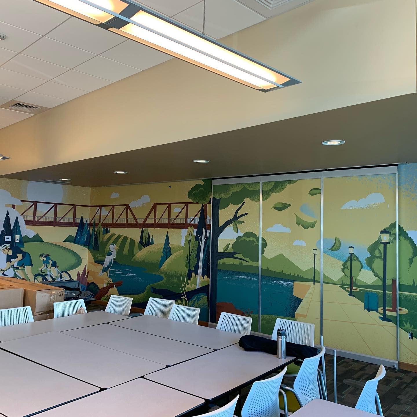 A conference room features a vibrant wall mural of a river, bridge, and landscape behind modular tables and chairs.