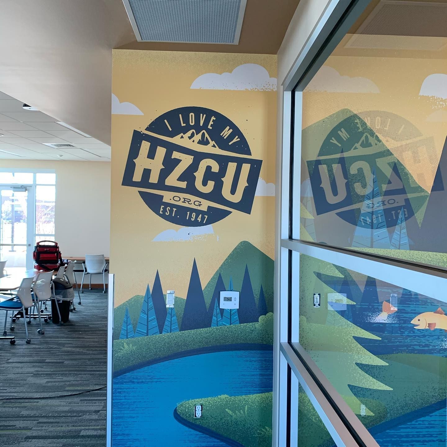 A wall mural depicting a mountain landscape with a blue lake, trees, and the HZCU logo in a modern office space.
