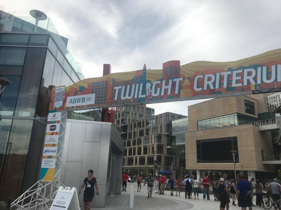 An overhead banner for the Twilight Criterium event hangs above a plaza with a modern building and people walking by.