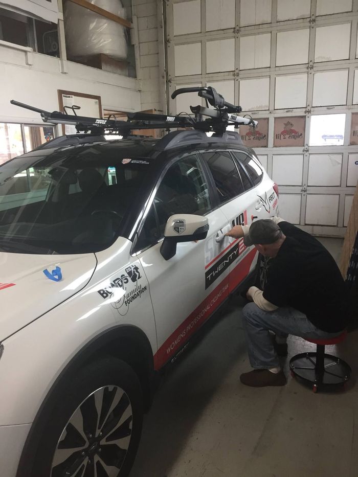 A person sits on a stool applying a decal to the side of a white vehicle inside a garage.