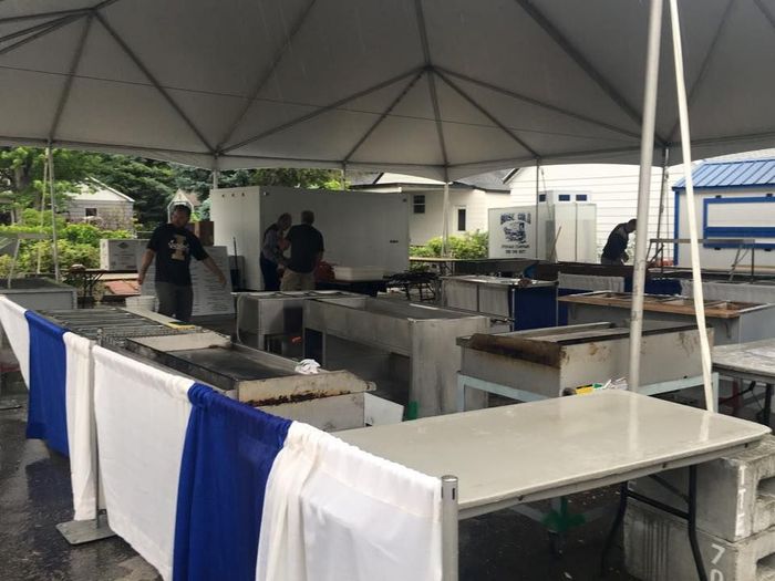 Several people work at outdoor food preparation stations under a large white event tent with blue and white skirted tables.