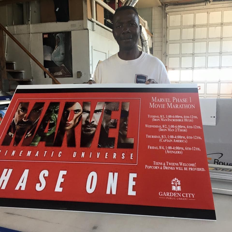 A person holding a large red sign promoting a Marvel Phase One movie marathon at Garden City Library.