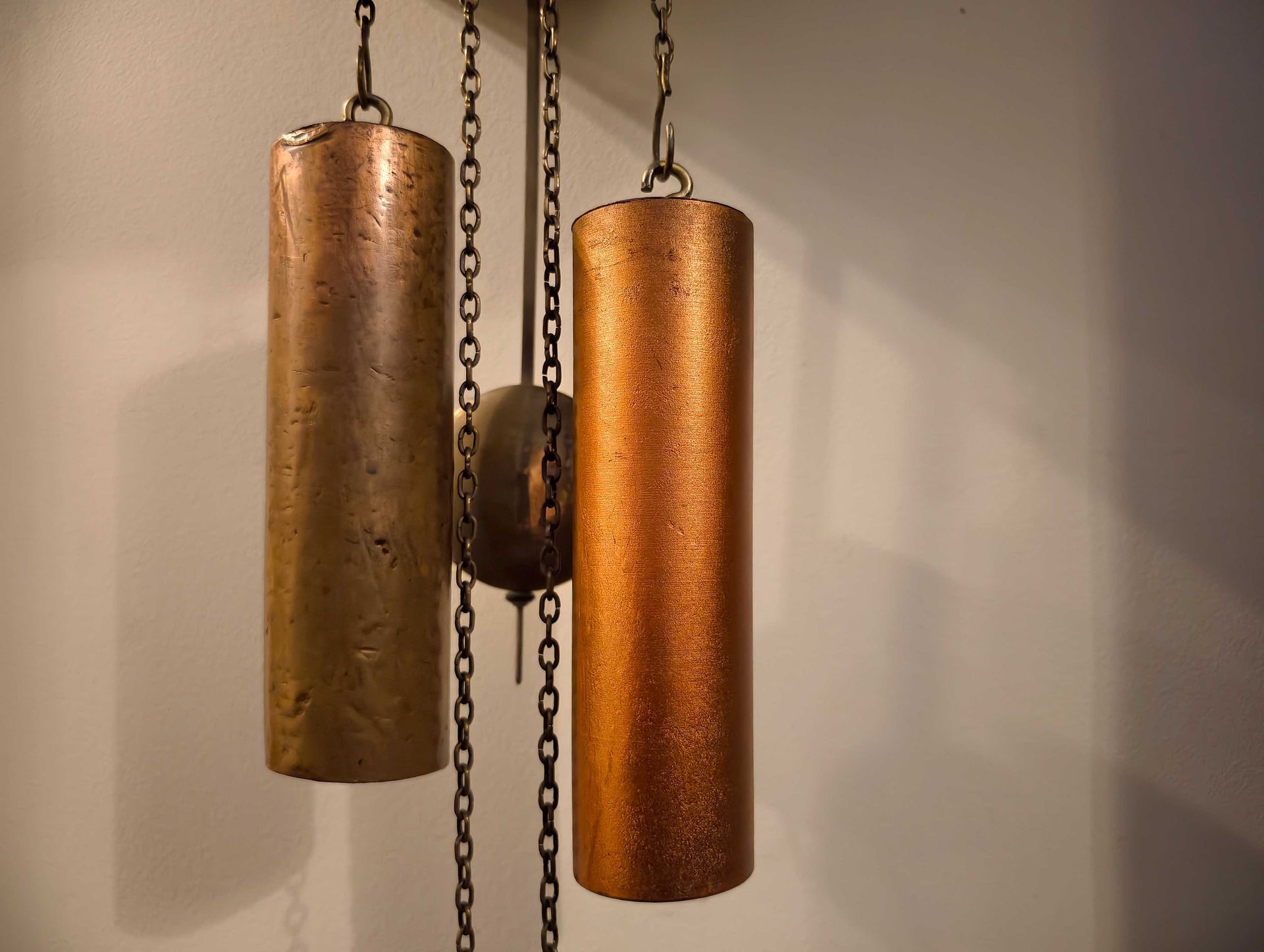 Deux poids d'horloge couleur cuivre suspendus à des chaînes contre un mur couleur crème.