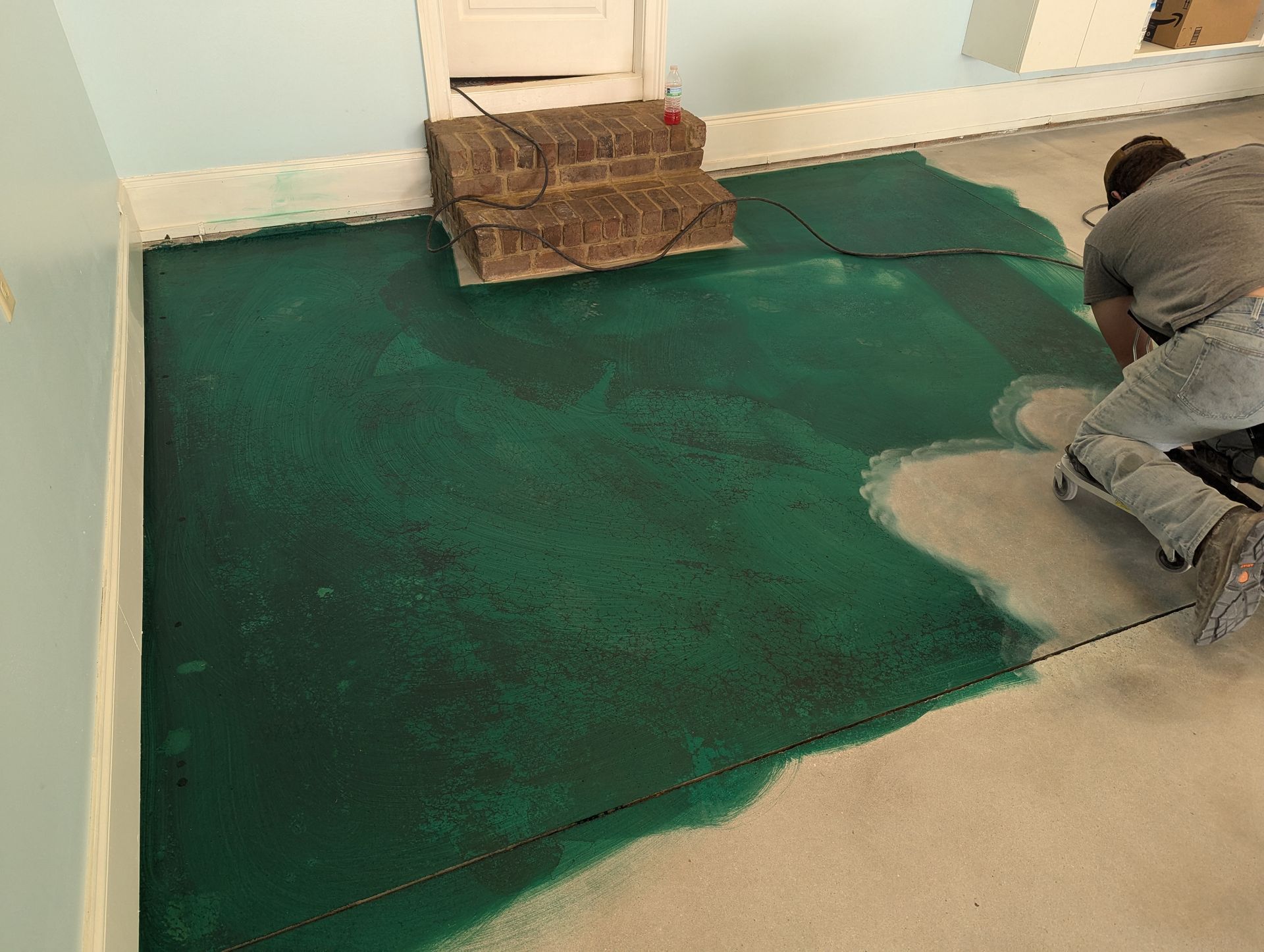 A man is kneeling on the floor painting a green rug.