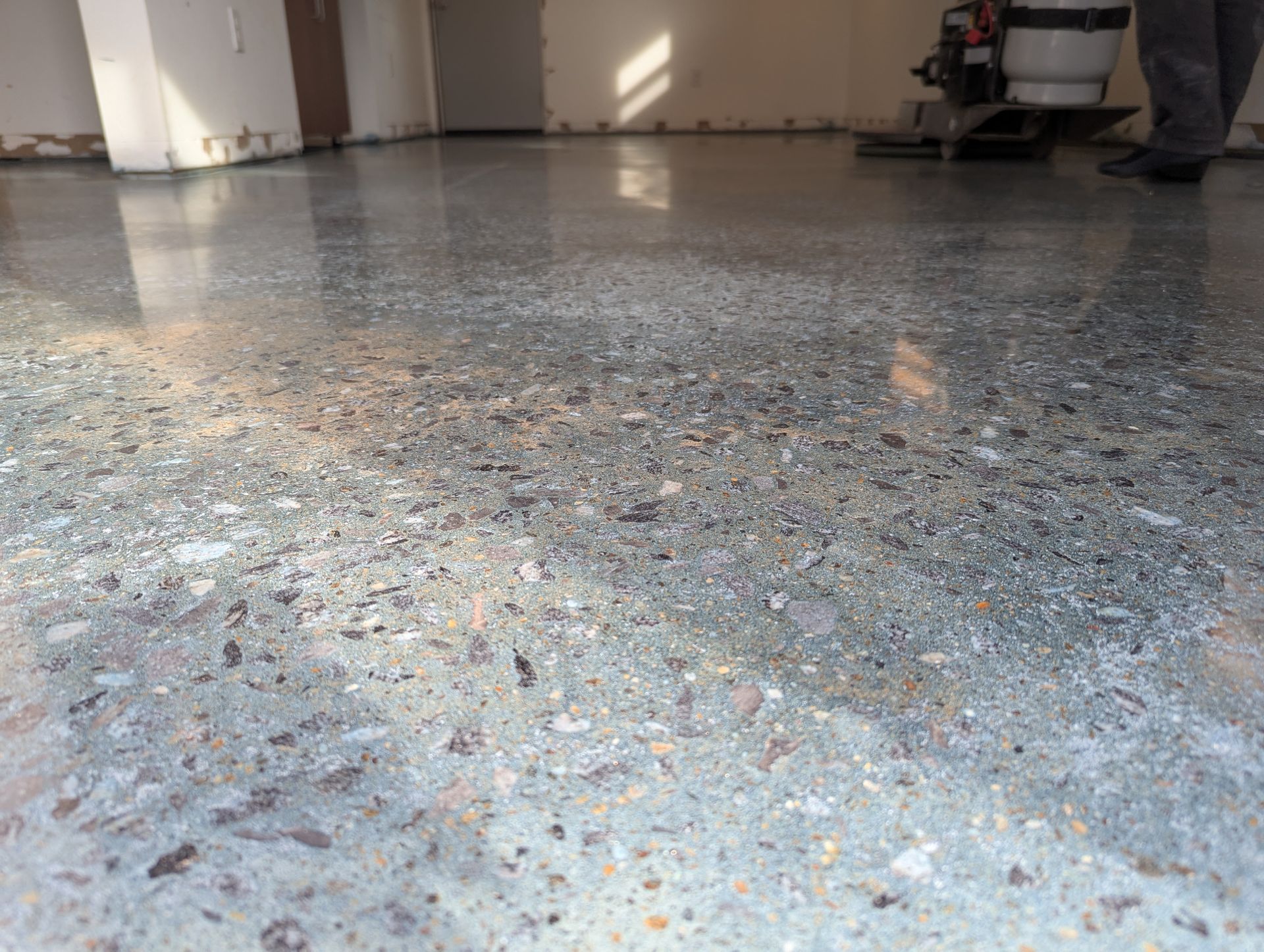 Glossy speckled concrete floor in a garage with sunlight reflections and a person’s legs at the edge