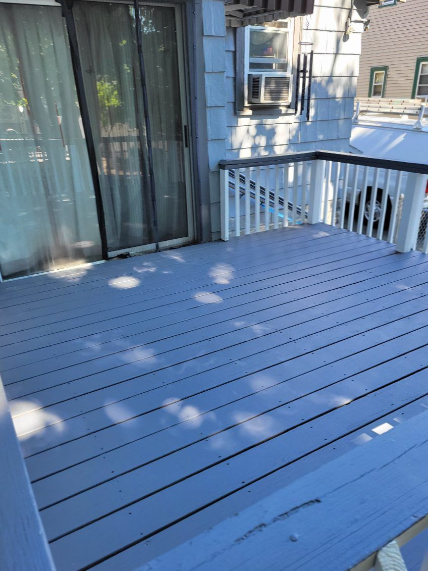 Blue wooden deck with white railing beside a sliding glass door and curtains, dappled in sunlight.