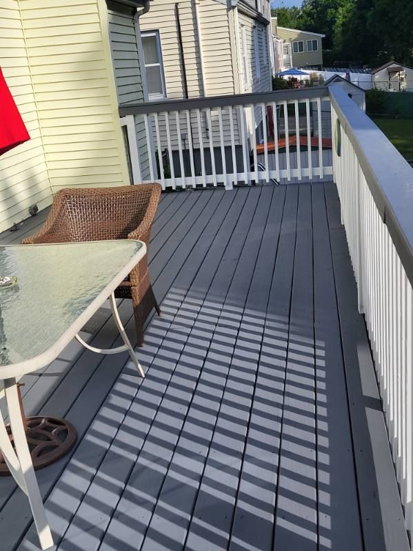 Sunlit gray deck with wicker chair, white railing, and striped shadows beside a house