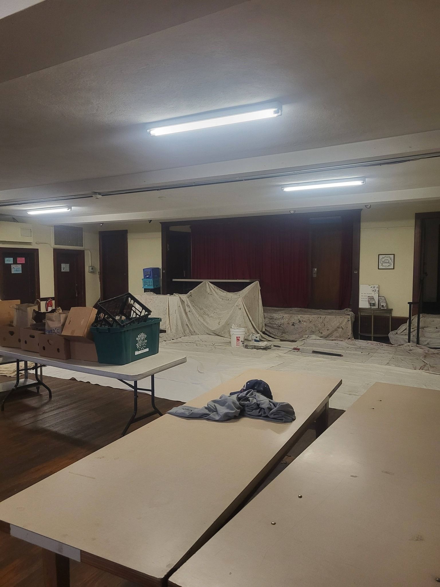 Unfinished hall with scattered boards, tarps, tables, and ladders under bright ceiling lights.