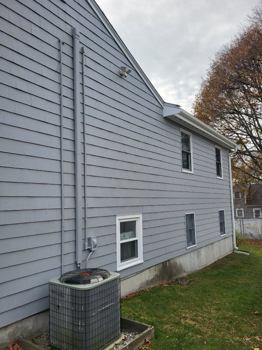 Gray house side with white trim, basement windows, and an air conditioner unit beside a grassy yard.