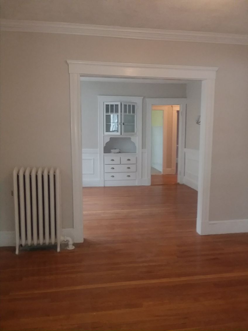 Empty room with hardwood floors, white walls, a radiator, and a doorway into a small built-in cabinet area.