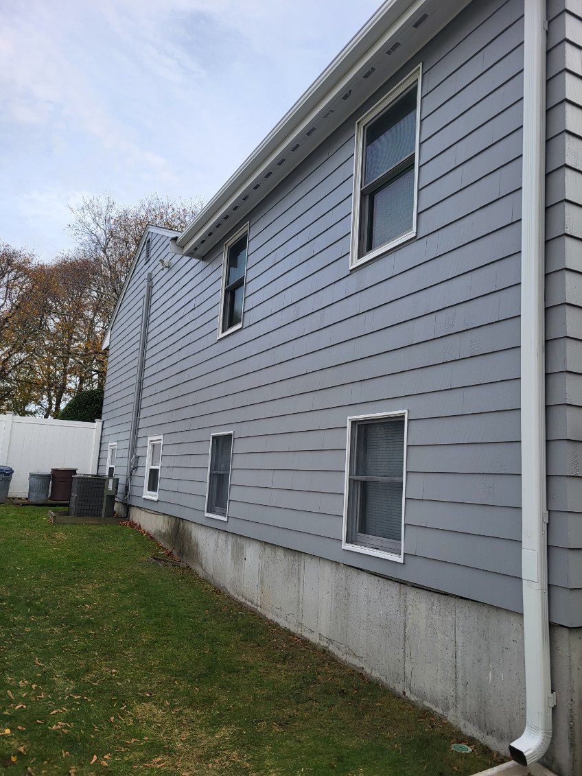 Side of a gray house with white trim, windows, and a concrete foundation beside a grassy yard.