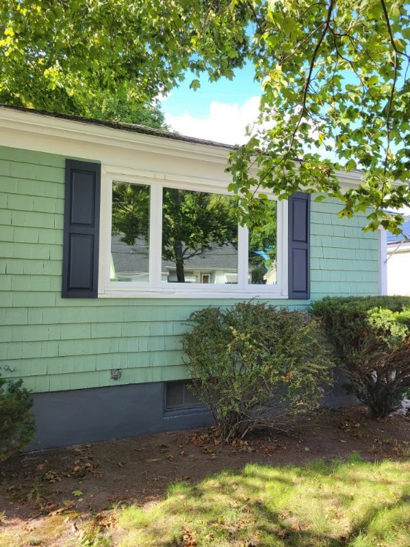 Light green house exterior with white-framed window, dark shutters, and sunlit trees overhead.