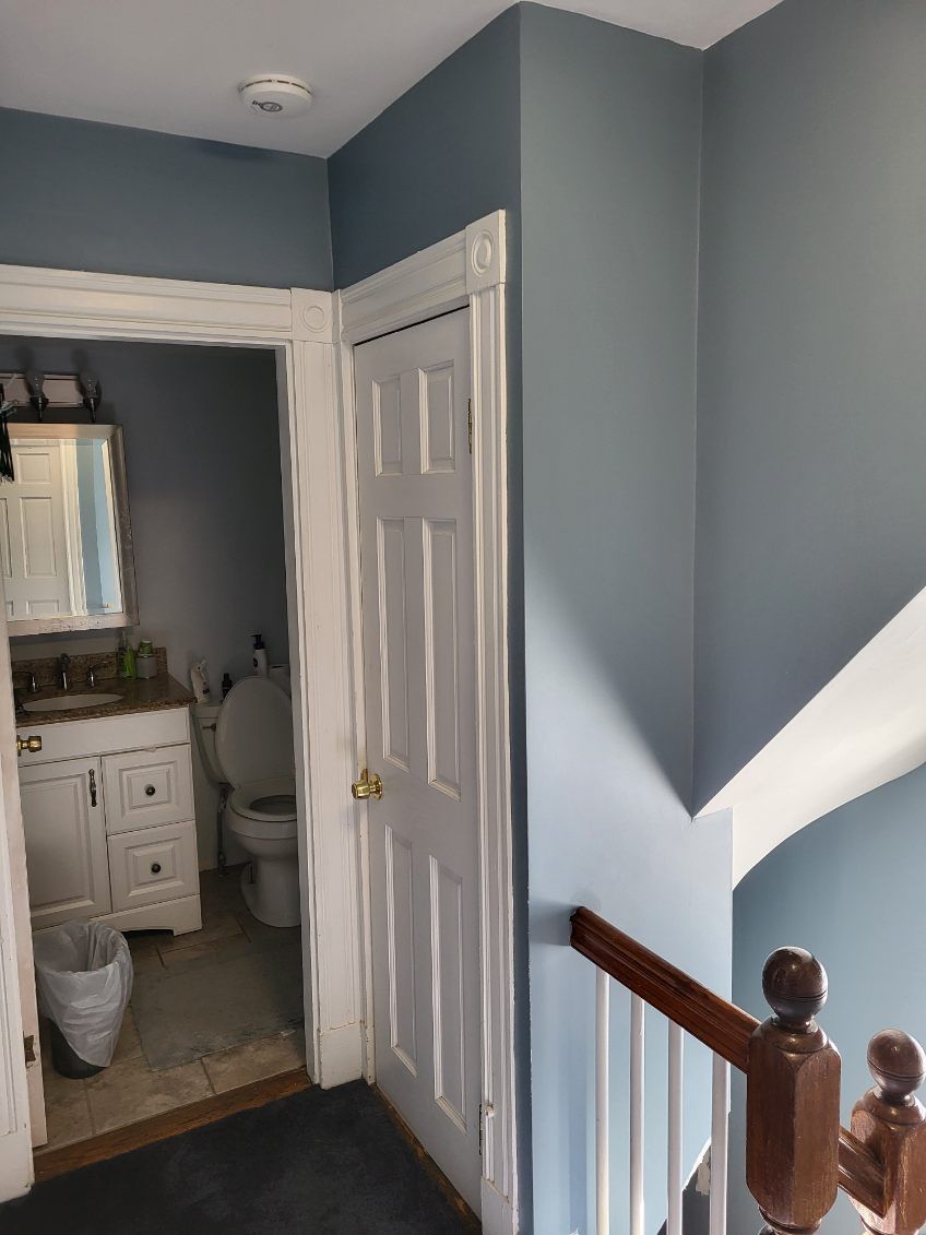 Hallway with blue walls, white doors, and a staircase railing, leading into a small bathroom.