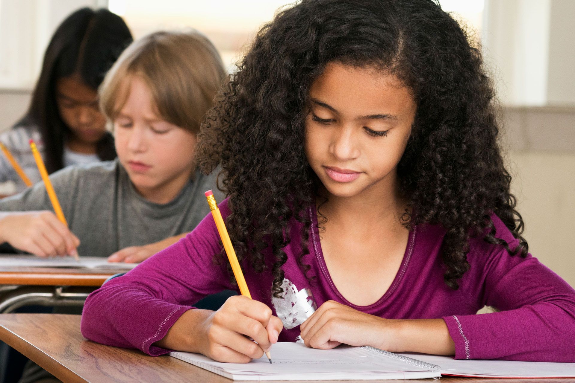A girl is writing in a notebook while sitting at a desk in a classroom.