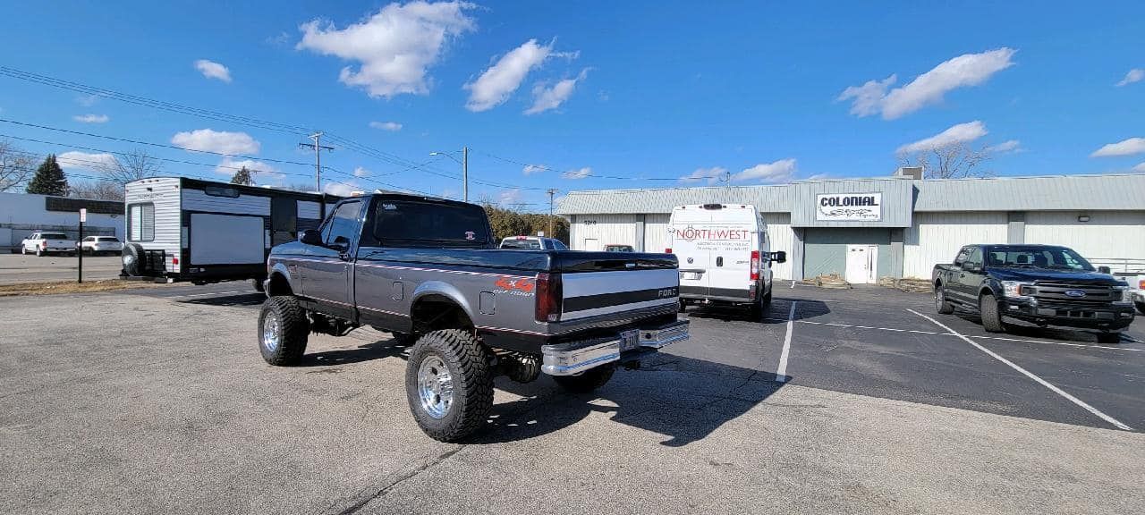 Lifted dark blue pickup truck parked in a gravel lot with RVs and cars under a bright sky
