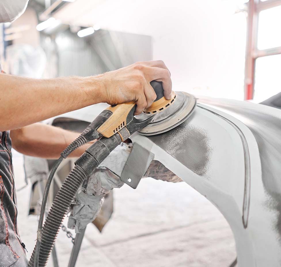 Hands sanding a car body with a power sander in a bright auto repair shop