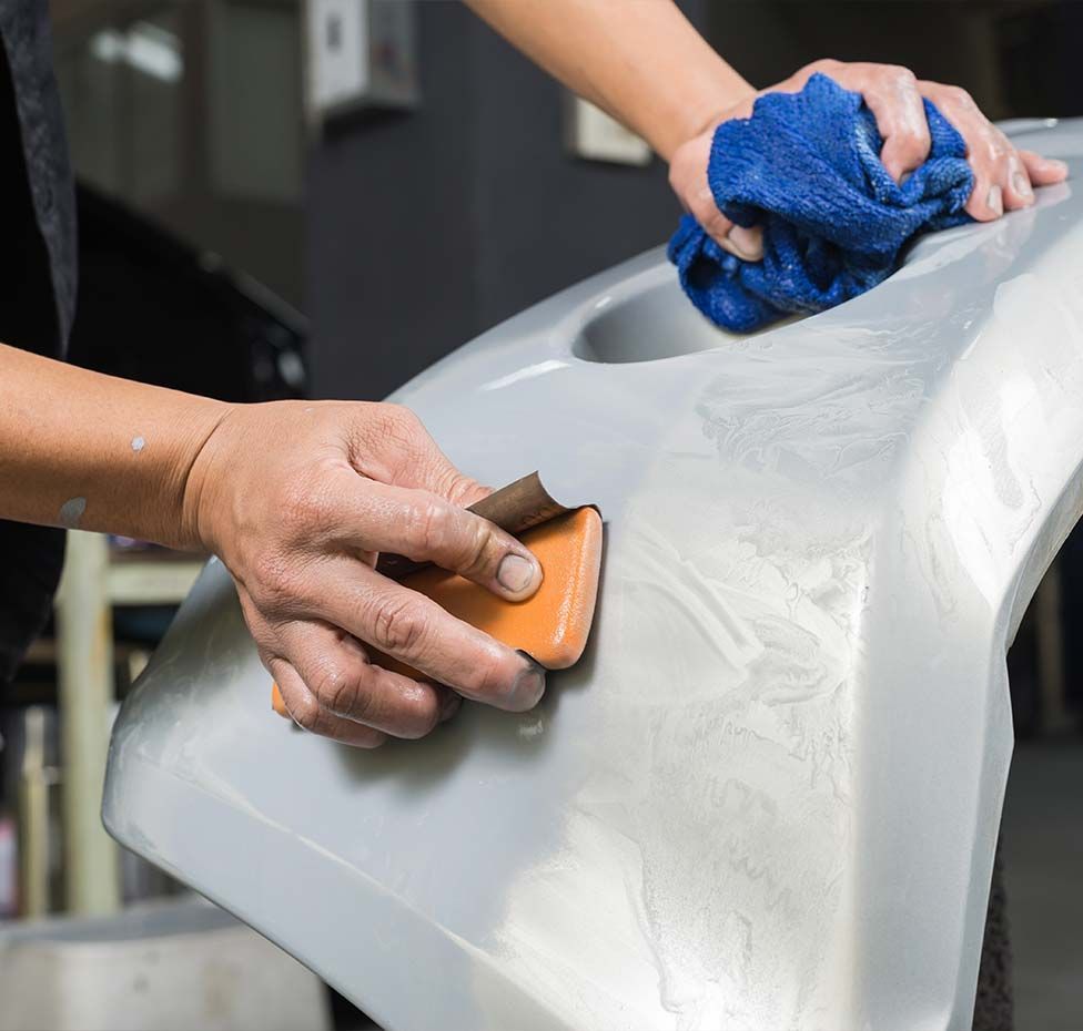 Hands sanding a white car body panel with a blue cloth in an auto repair shop