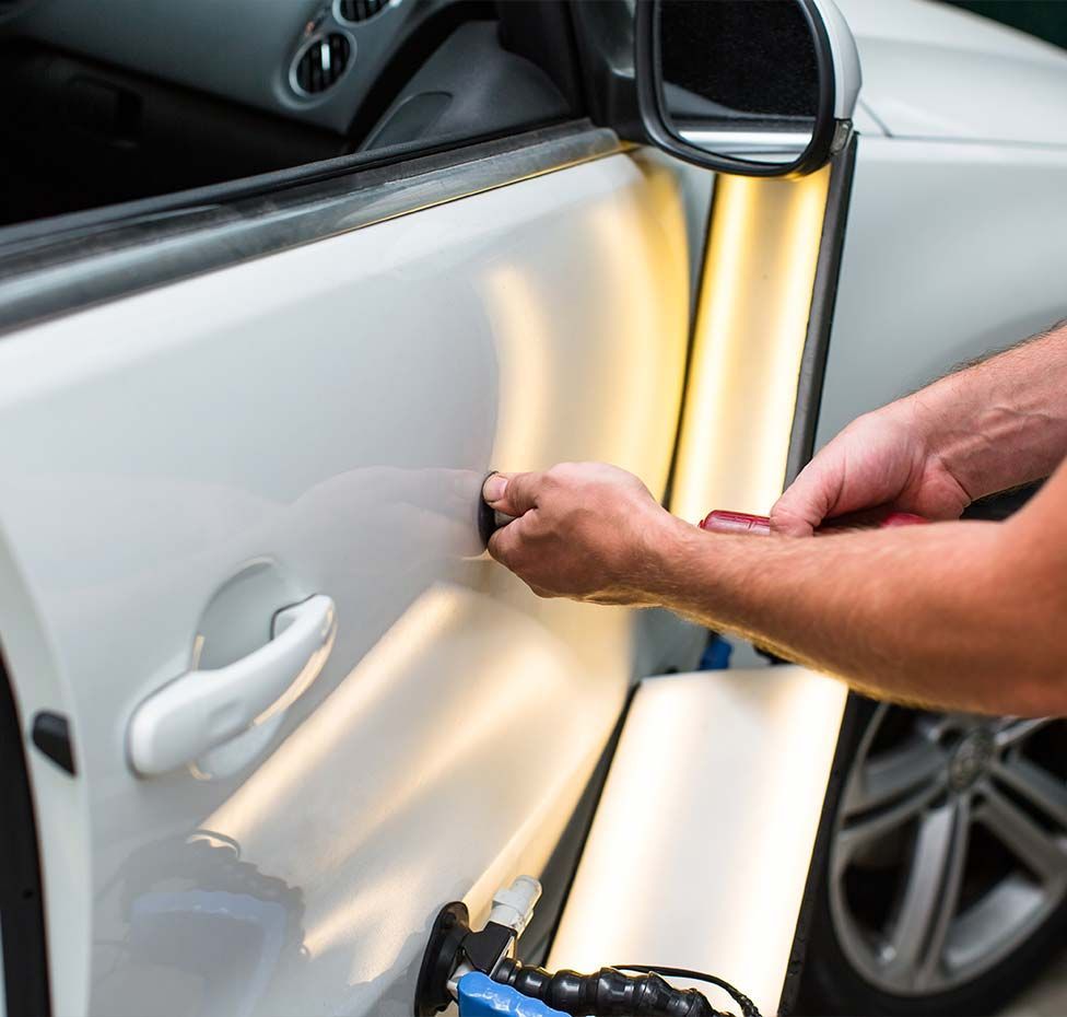 Person repairing a white car door with a metal tool and clamp in a garage