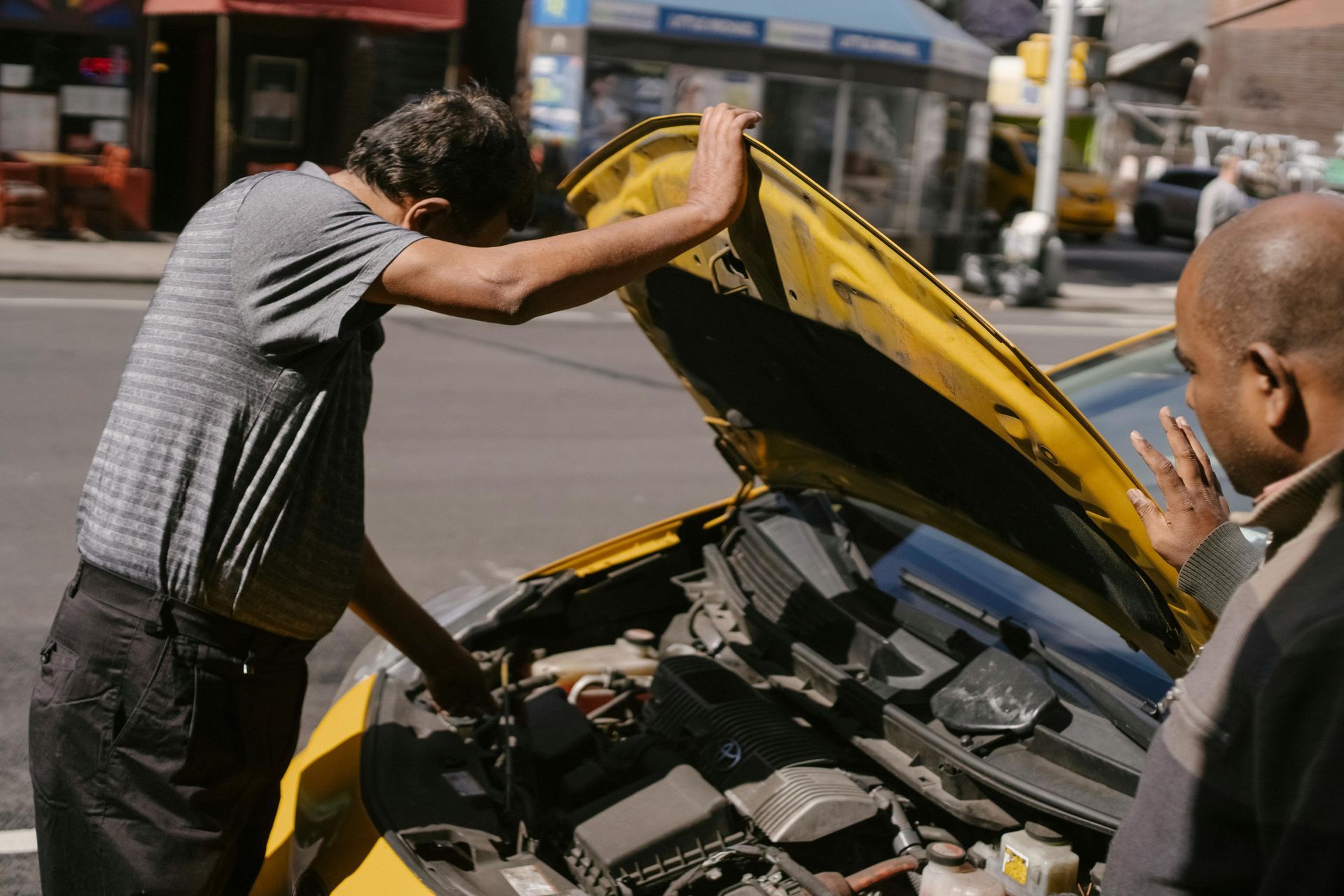 Two people inspect a yellow car with its hood open on a city street.