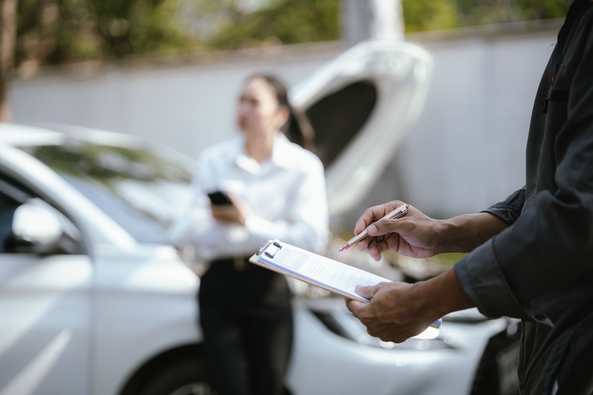 Person holding a clipboard beside a white car while another stands near the open hood