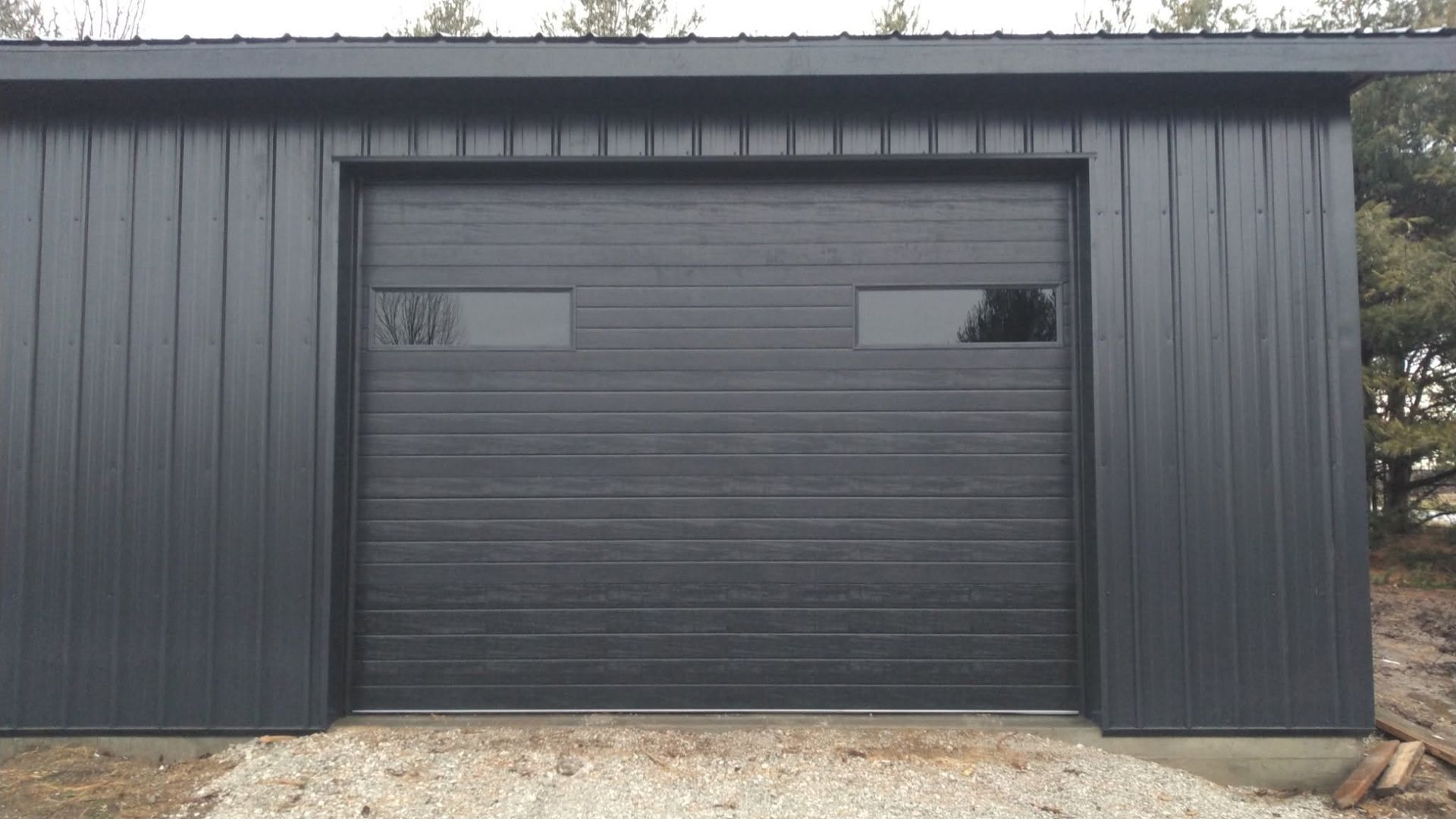 Dark gray metal garage with a large roll-up door and two small windows, centered on a gravel drive.