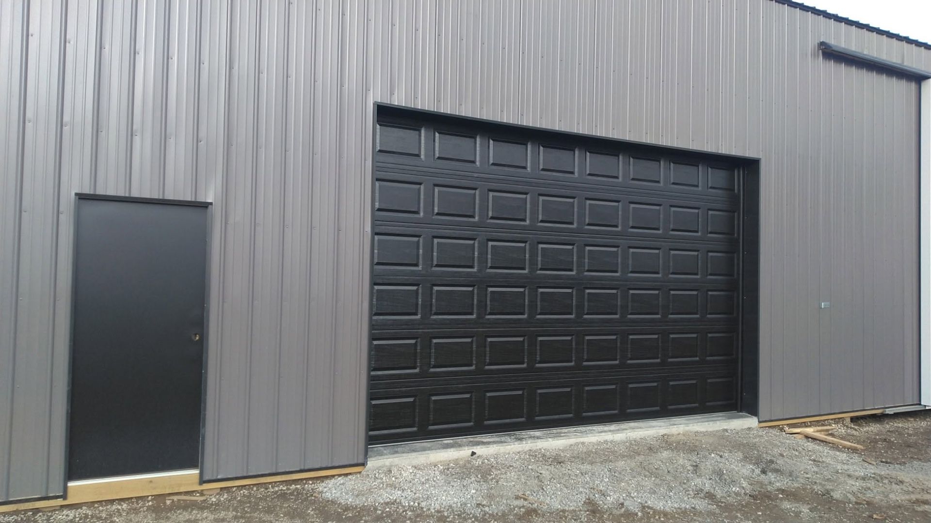 Gray industrial building with a large black garage door and a small side door on gravel ground