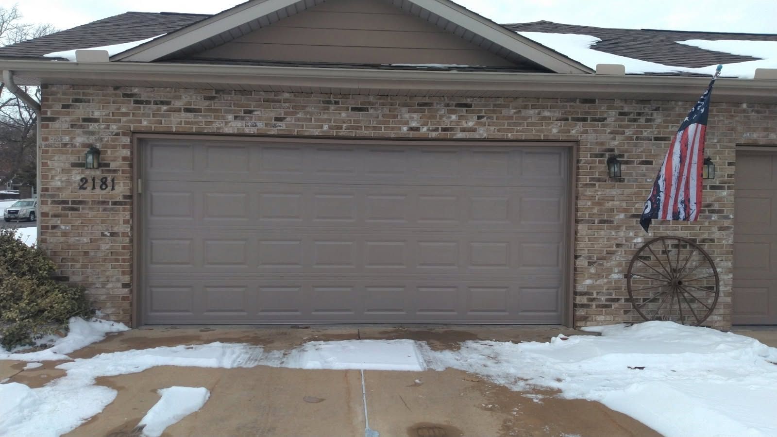 Snowy suburban house with a closed brown garage door and an American flag by the entryway