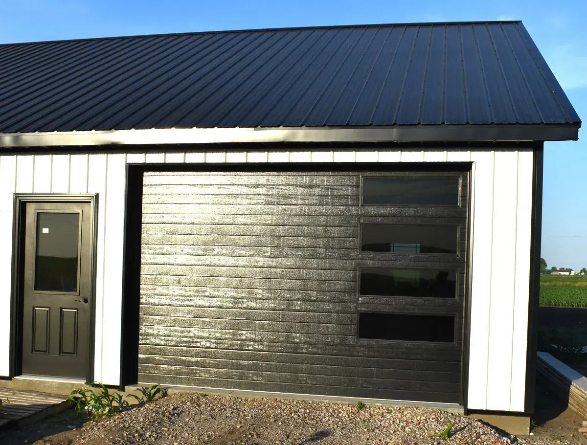 Small white building with a black metal roof and large dark garage door on a gravel lot.