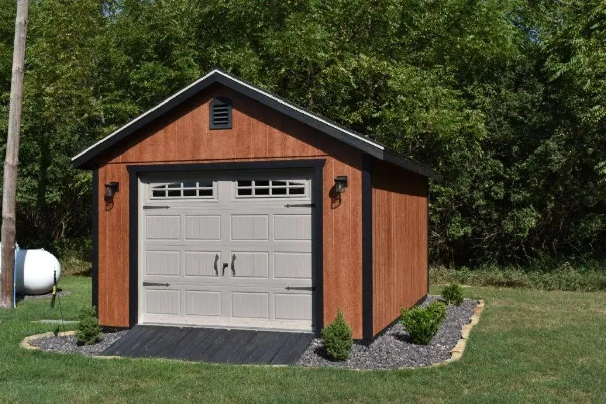 Small brown detached garage with a gray door and black roof in a grassy yard.