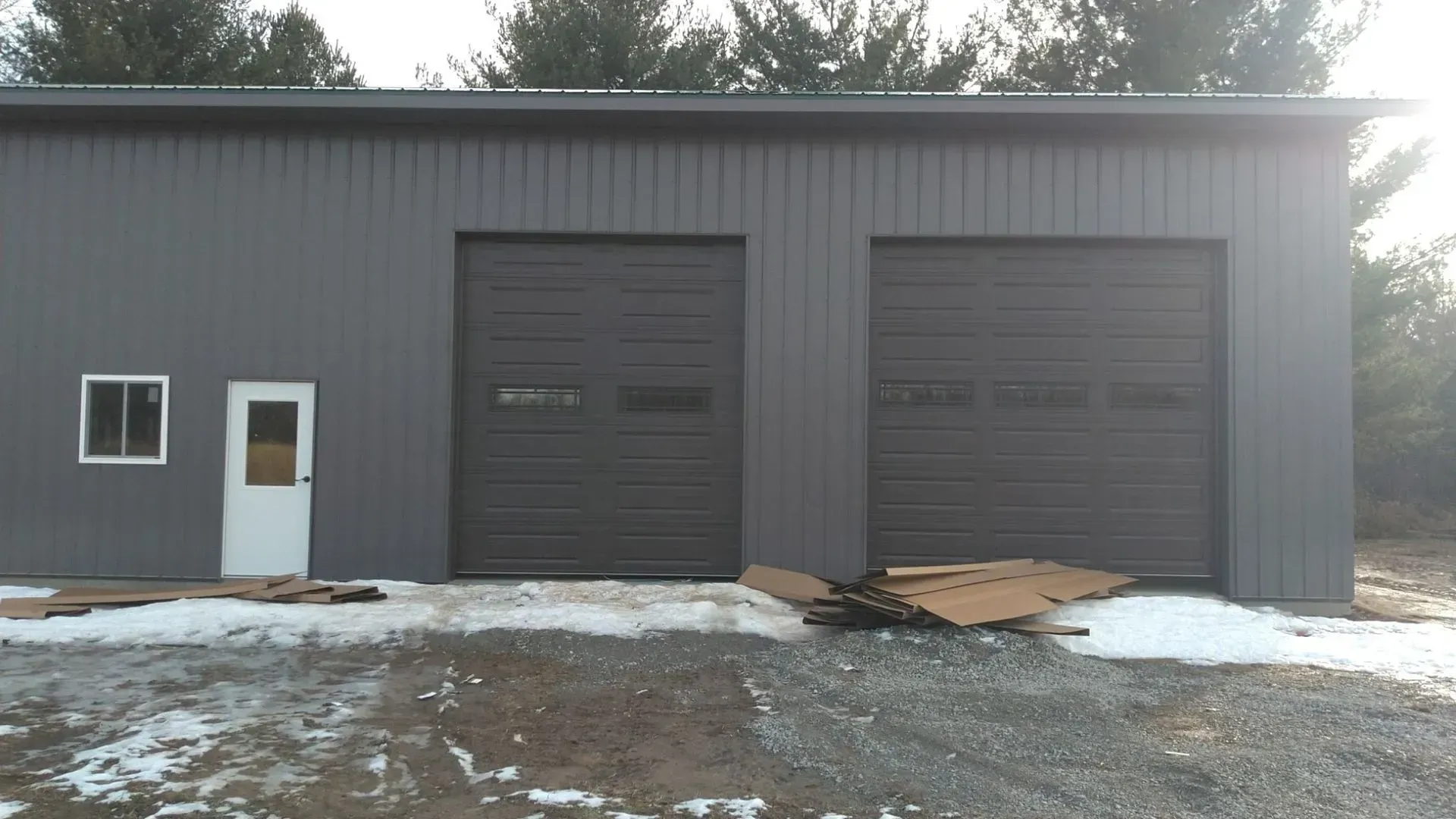 Gray metal garage building with two large bay doors, a side door, and snow on the ground.