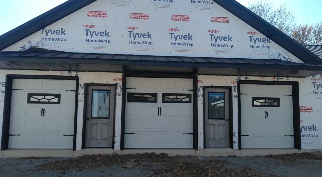 Front view of a house under construction with white garage doors and Tyvek-wrapped siding