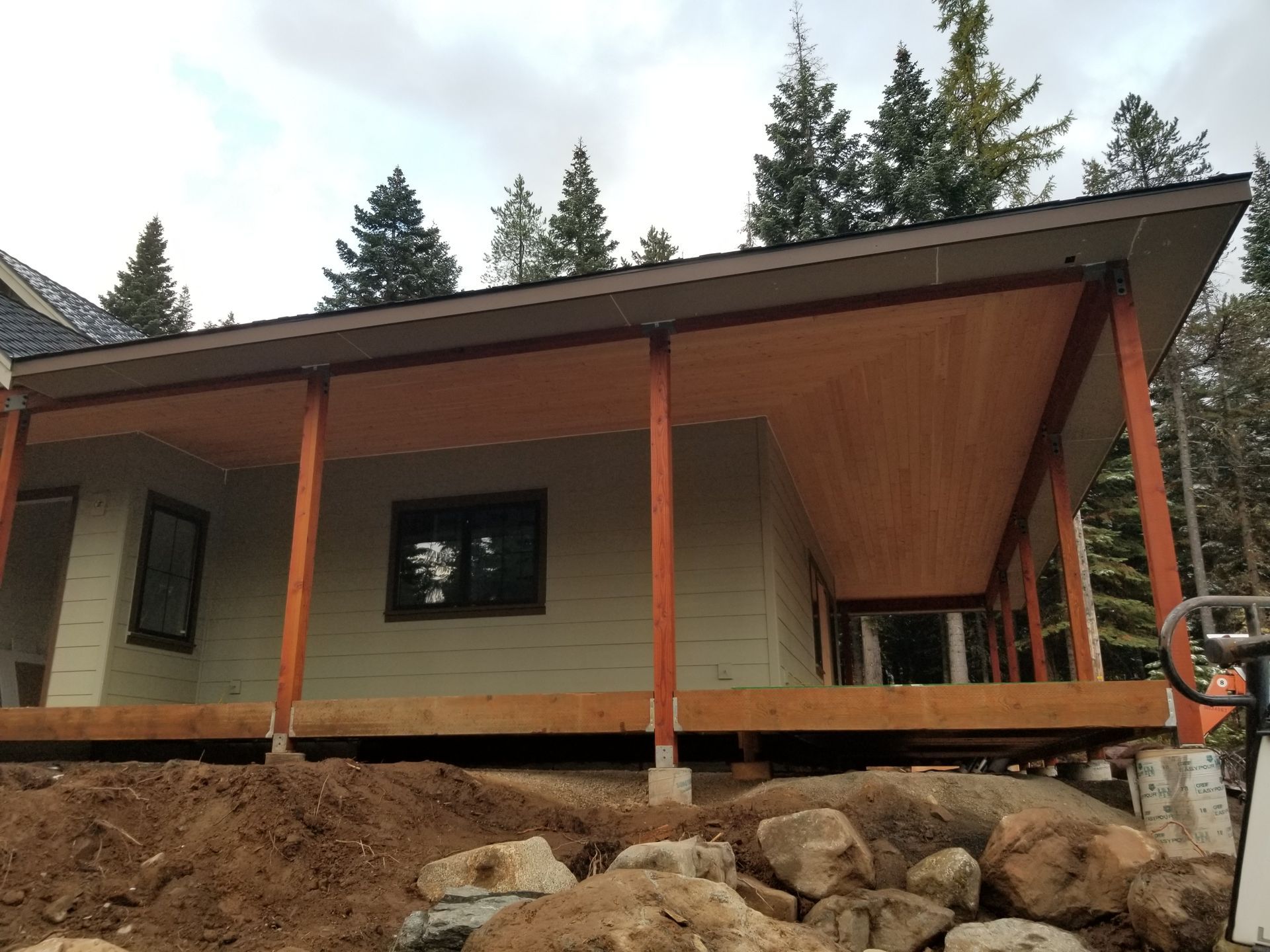 Modern house under construction with a sloped roof, wooden posts, stone foundation, and forested background