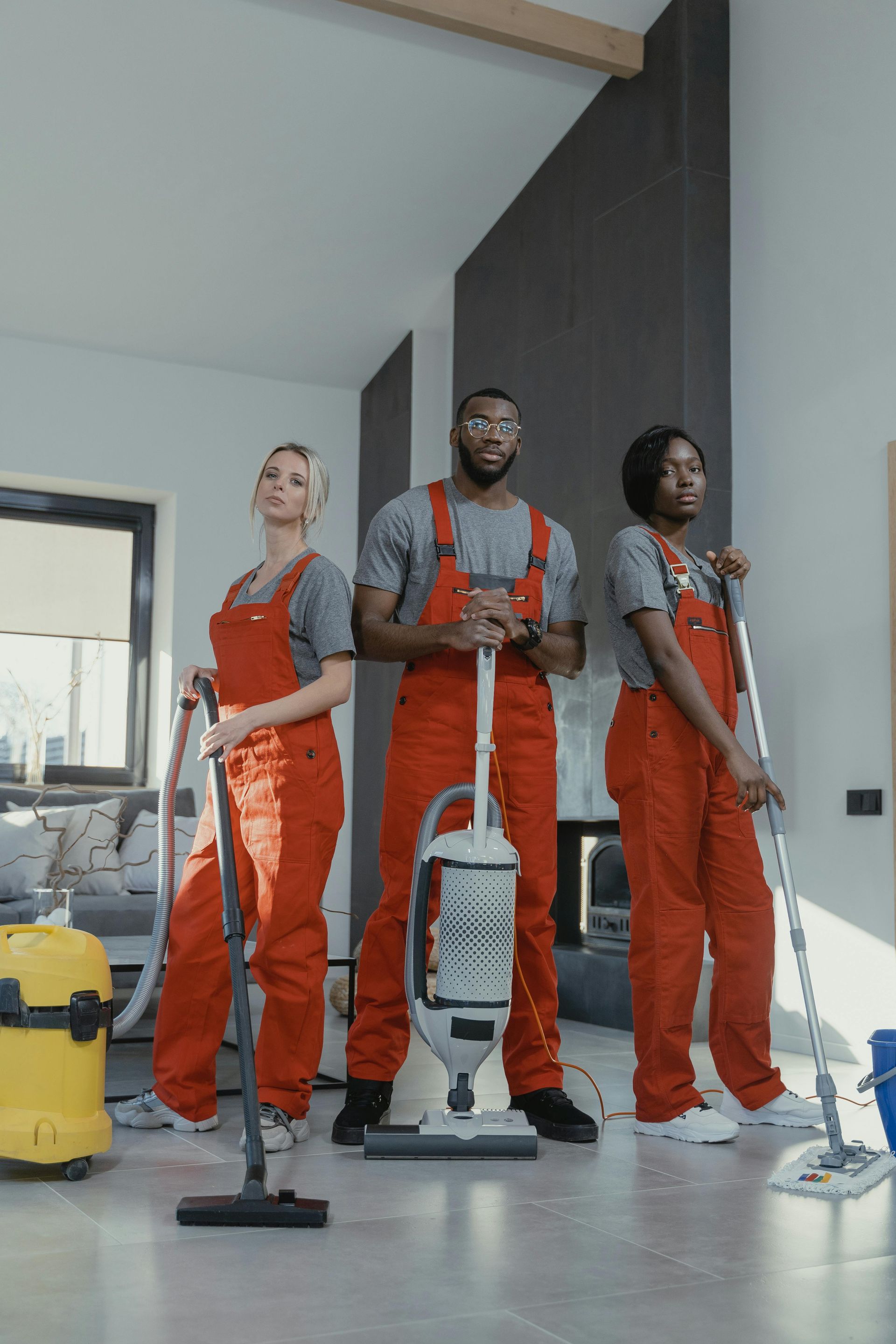 Three cleaners in orange coveralls with mops and vacuum in a bright hallway