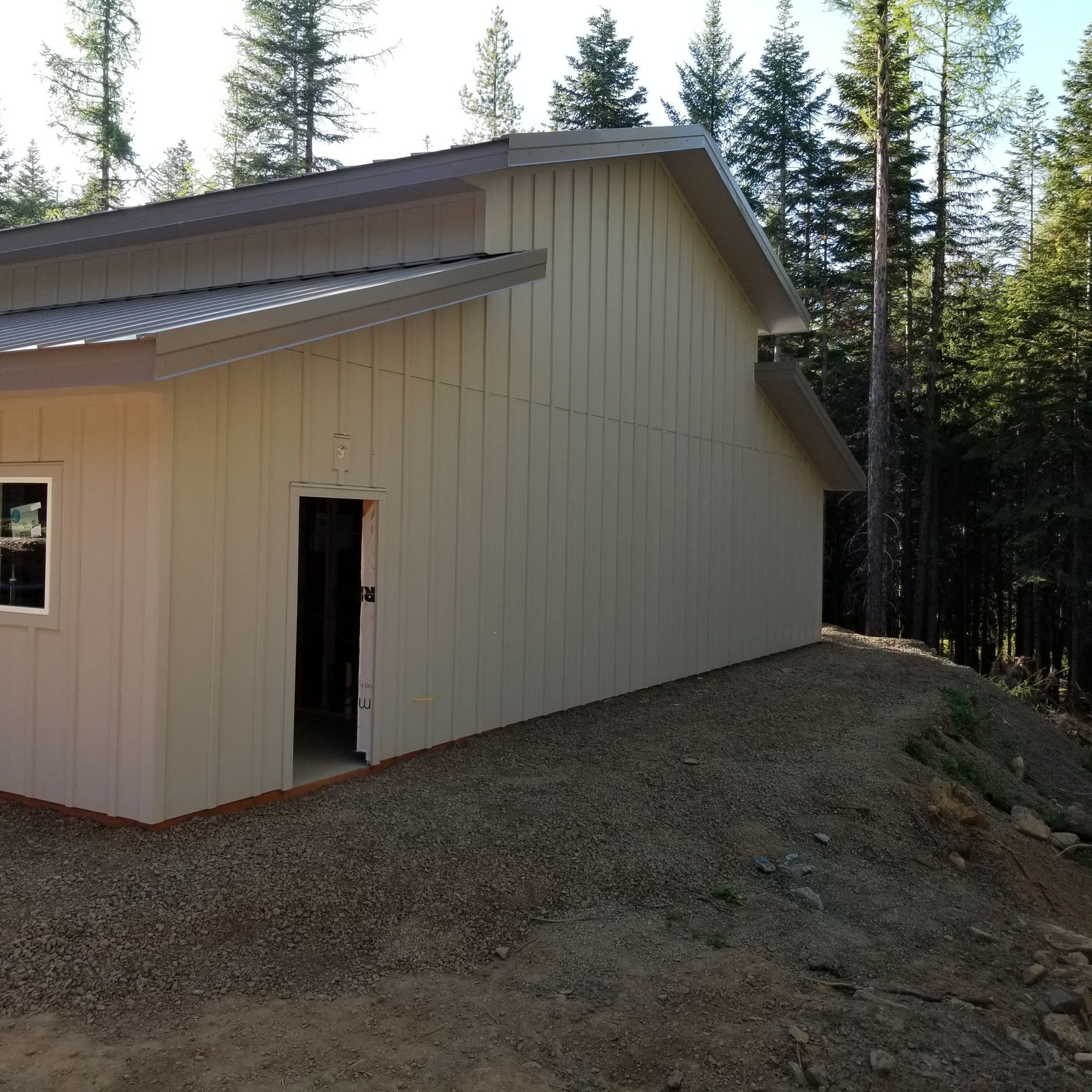 Beige metal building with open doorway beside a gravel slope and tall pine trees