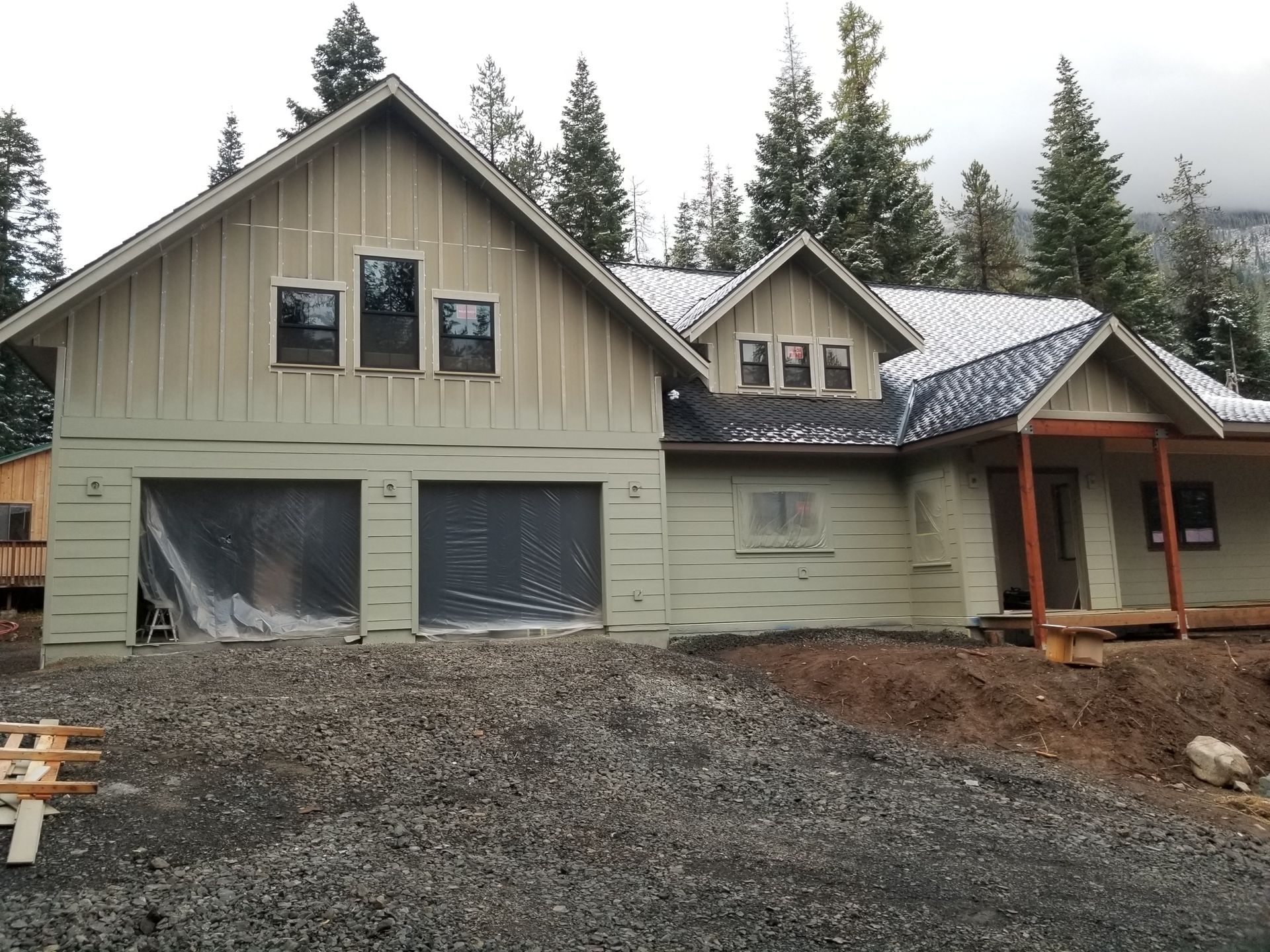 New beige house under construction with gravel driveway and tall trees in the background