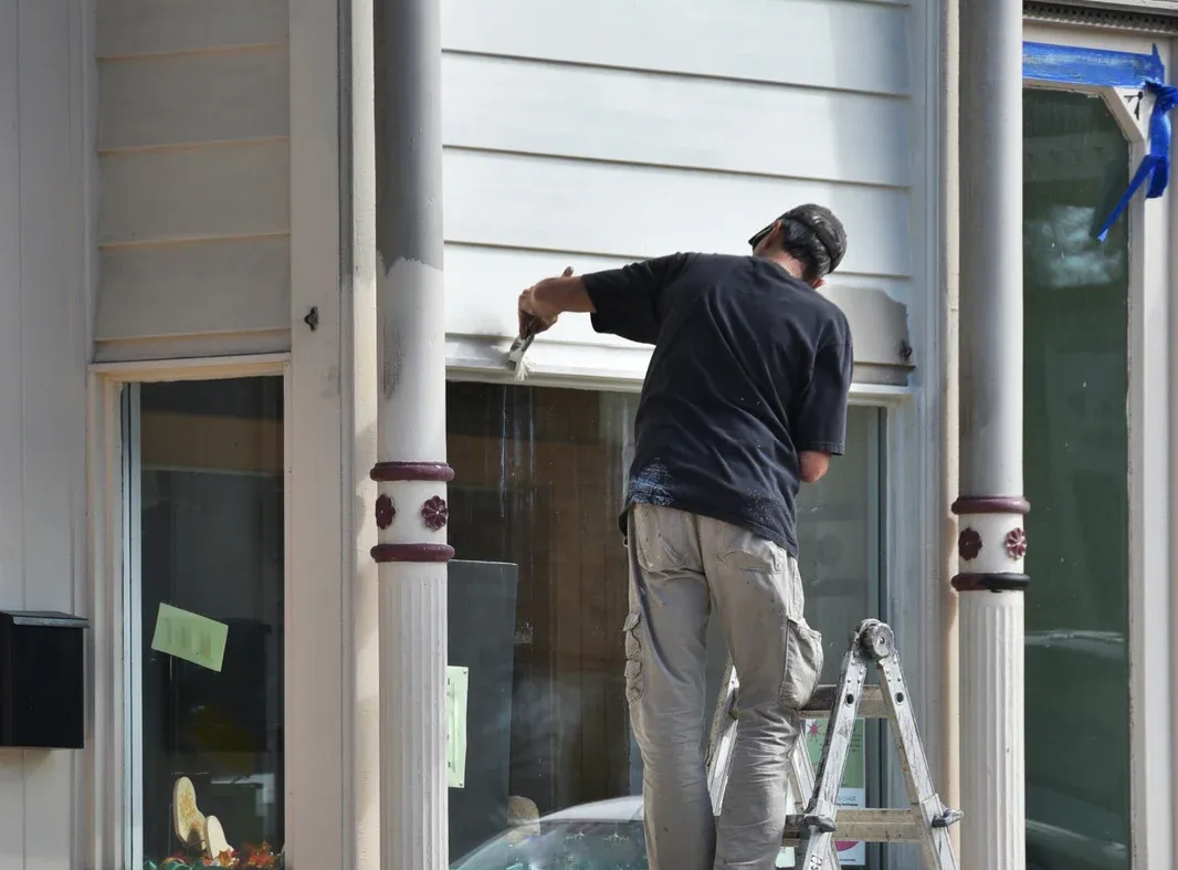 Worker on a ladder painting a storefront window frame beside a sidewalk