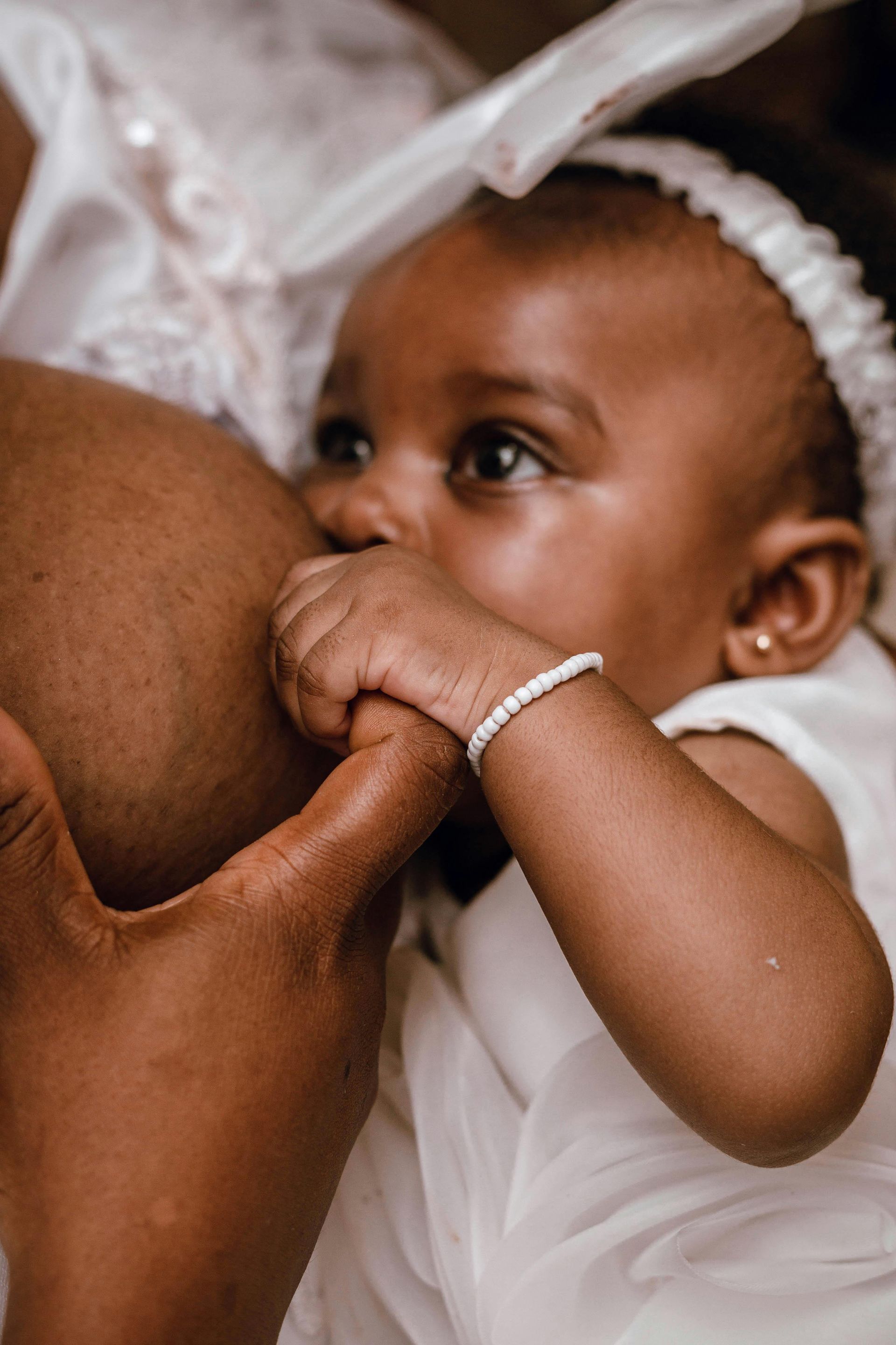 Baby nursing, wearing a white headband and bracelet, holding onto mother's finger.