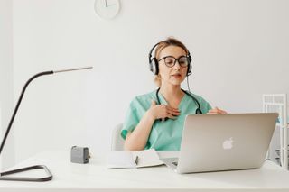 Medical professional in scrubs and headphones on a video call, laptop on white desk.