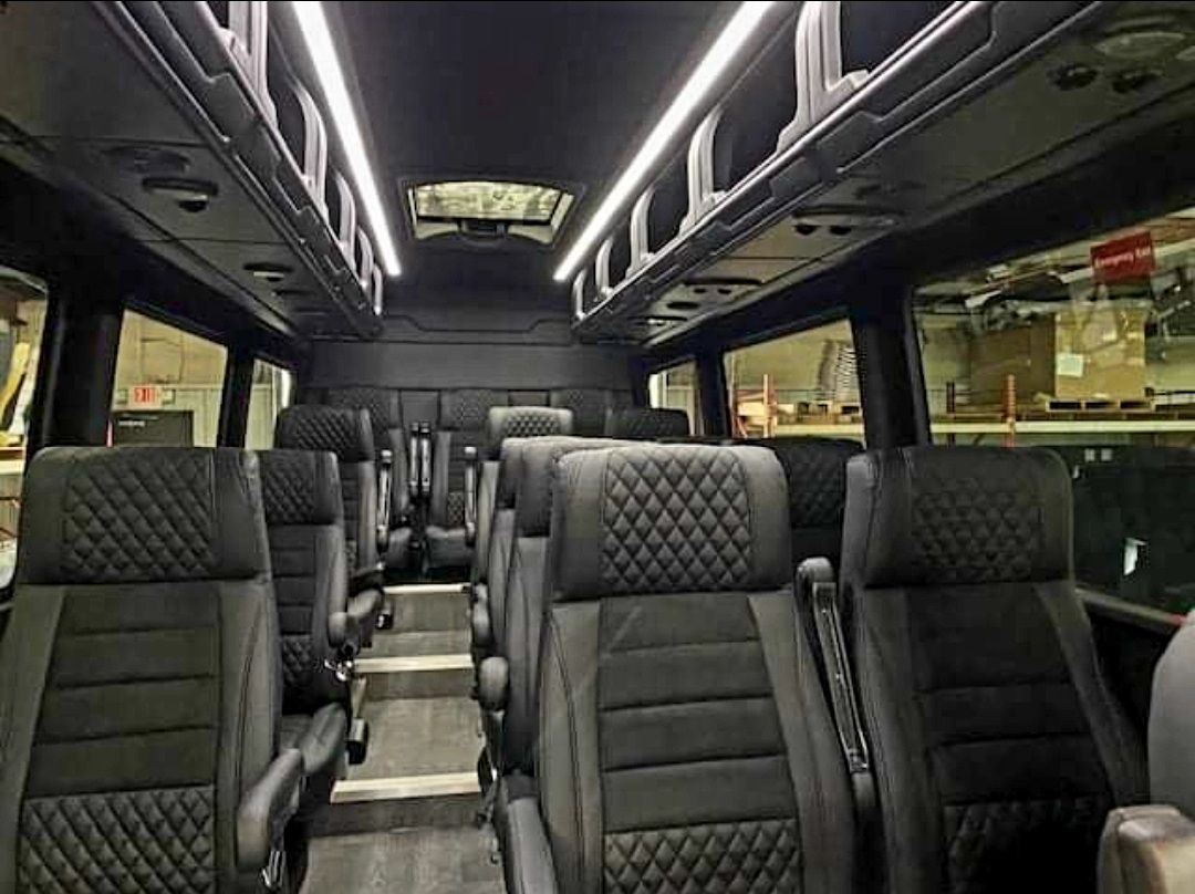 Interior of a passenger van with rows of black seats, overhead storage, and ceiling lights.