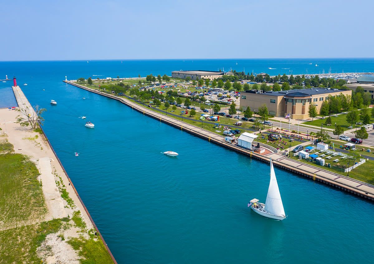 Sailboat on blue water waterway, with buildings and park on either side.
