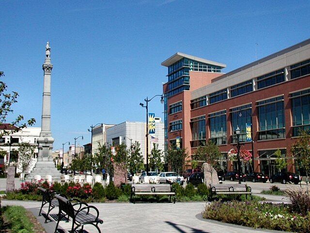 A city square with a tall monument, benches, and buildings under a blue sky.