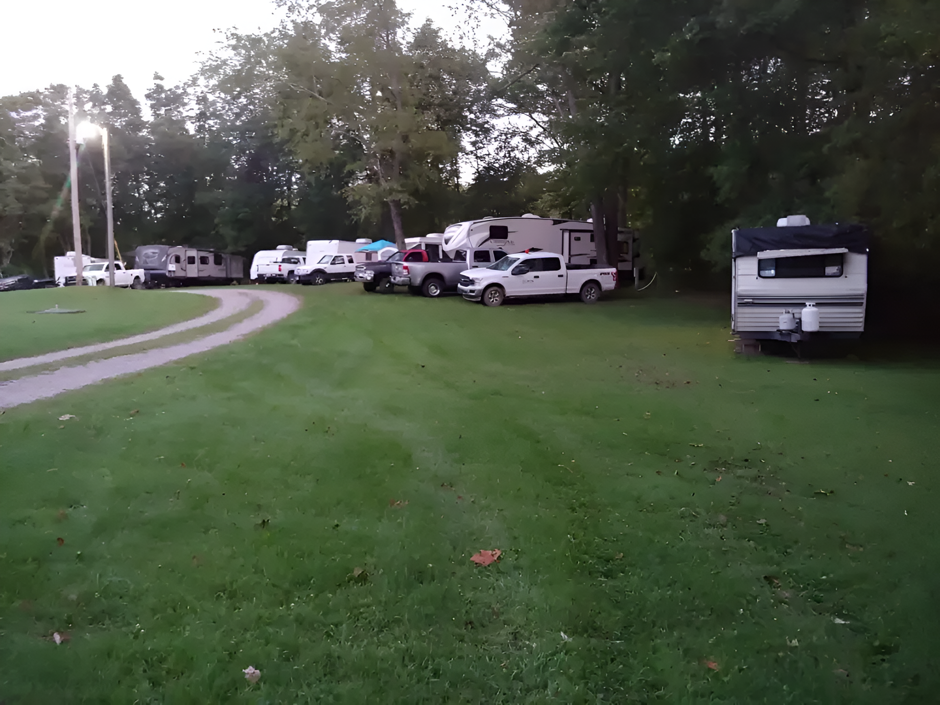 Campground with RVs and trucks parked on grassy lawn, trees in the background. Cloudy sky.