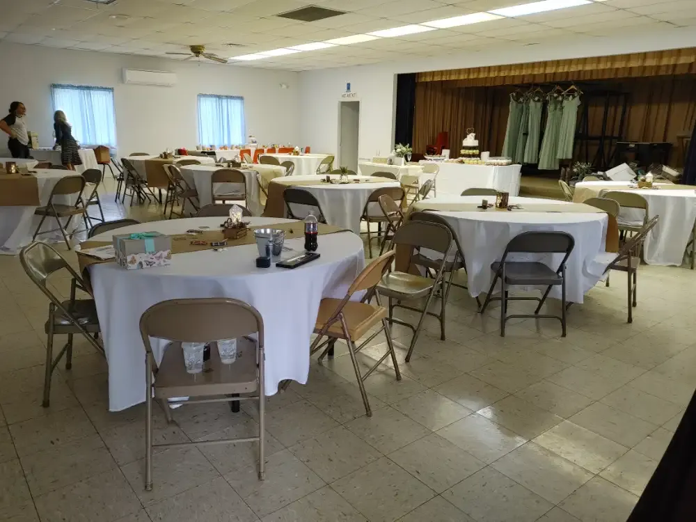 Wedding reception setup in a brightly lit hall, round tables draped with white cloths, brown chairs.