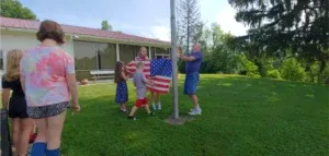 Family raising an American flag on a pole in a green yard.
