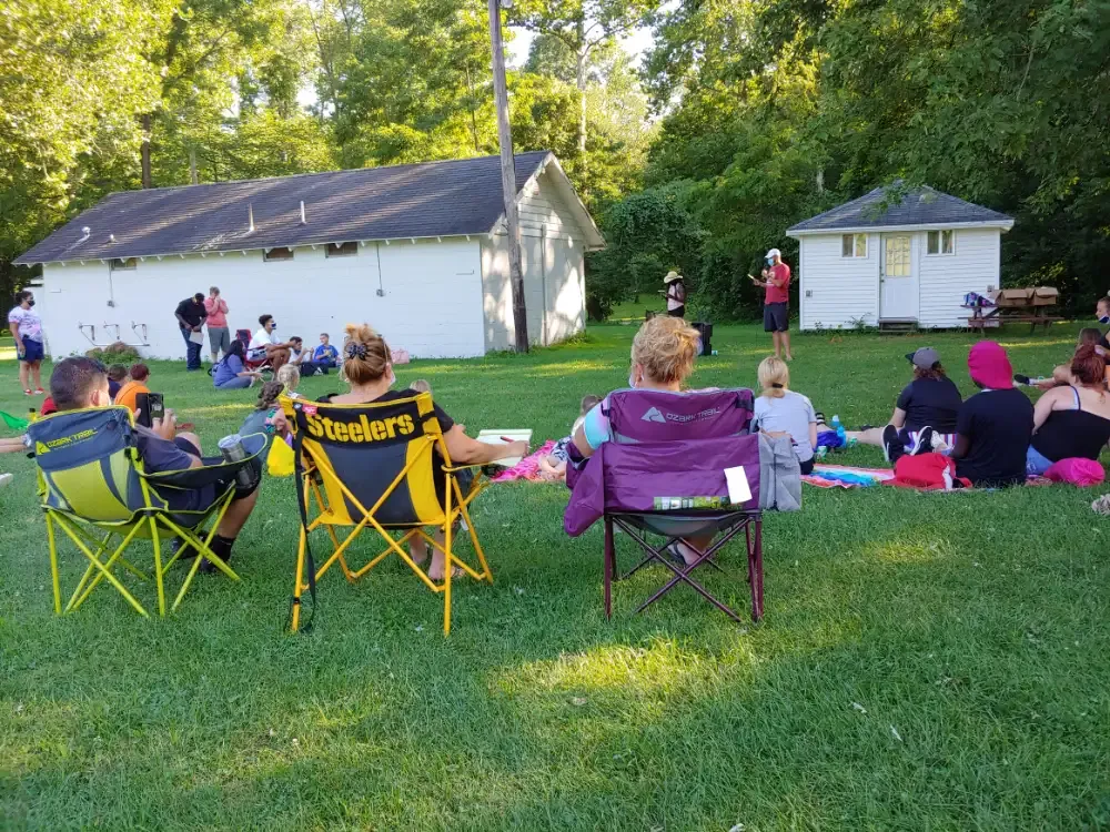 People gathered outdoors on the grass, watching a presentation near white buildings.